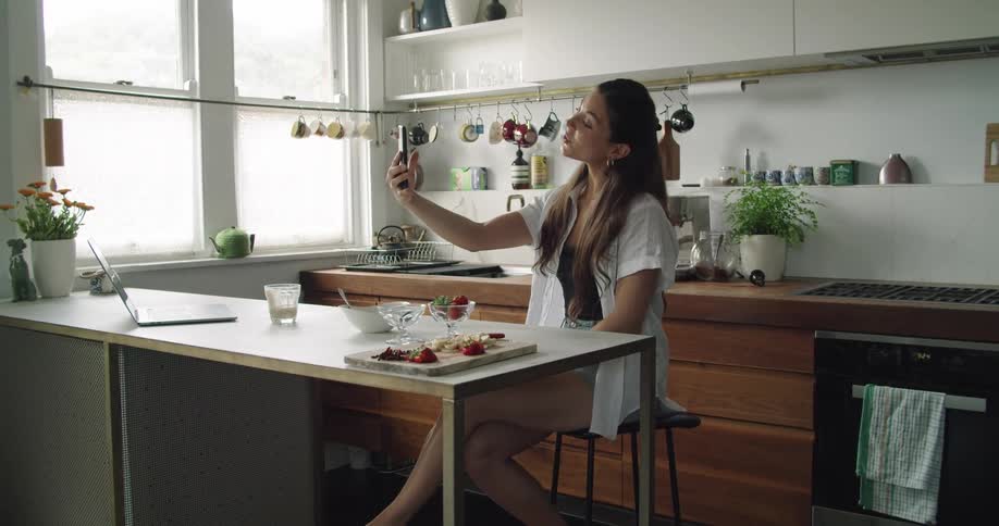 A woman holds out her phone and waves to the screen as she sits in a kitchen