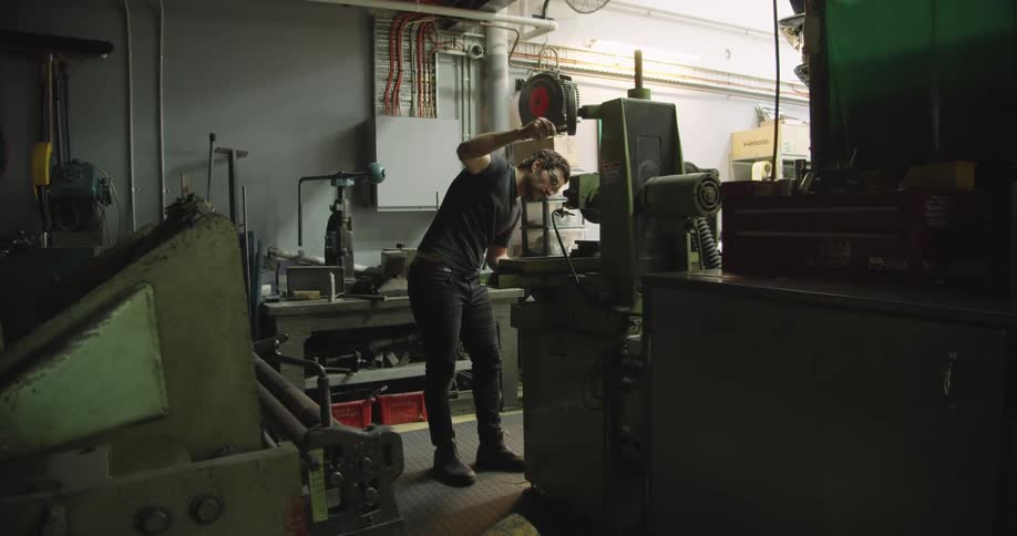 A man stands in a large garage or workshop with machines turning the handle on something above him