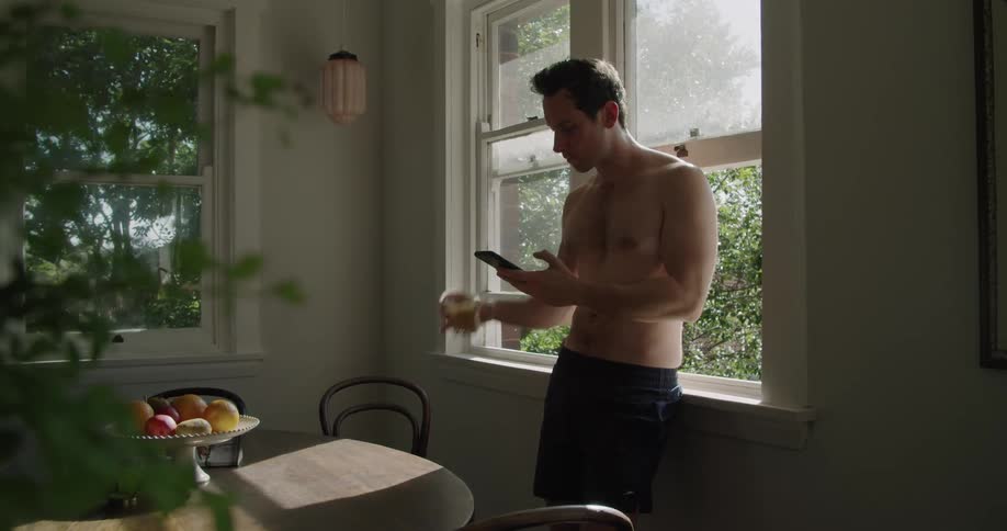 A shirtless young man standing while leaning against the sill of an open window drinks from a glass while using his smartphone