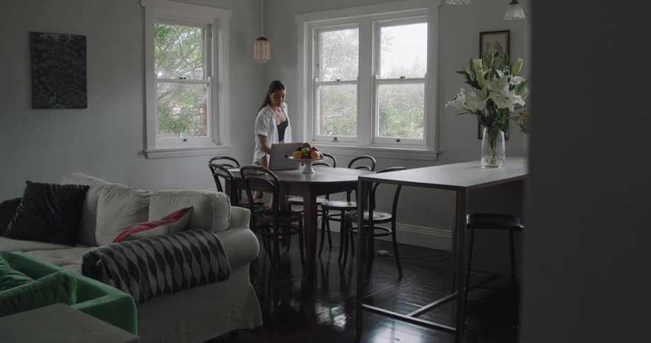 Woman picks up a mug off of a counter top and walks over to a dining room table and sits in front of a laptop computer during the day