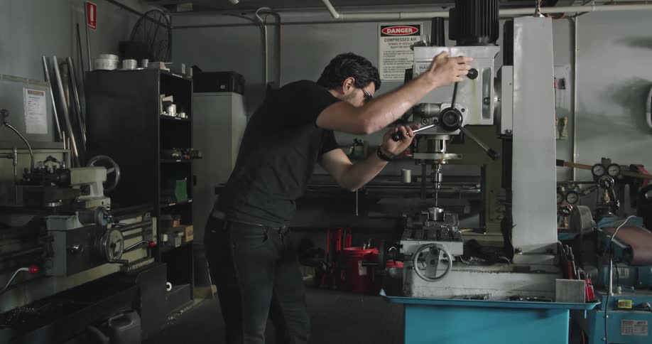 Man with safety glasses in a machine shop preparing a drill press to drill into a piece of metal