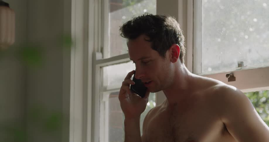 Young fit shirtless man answers a phone call on a smartphone at home