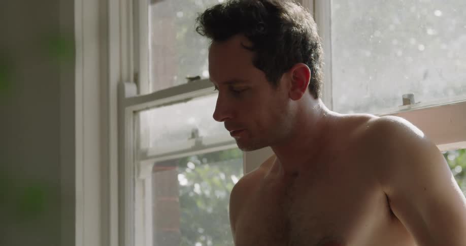 A bare chested man stands inside against a window drinking from a glass
