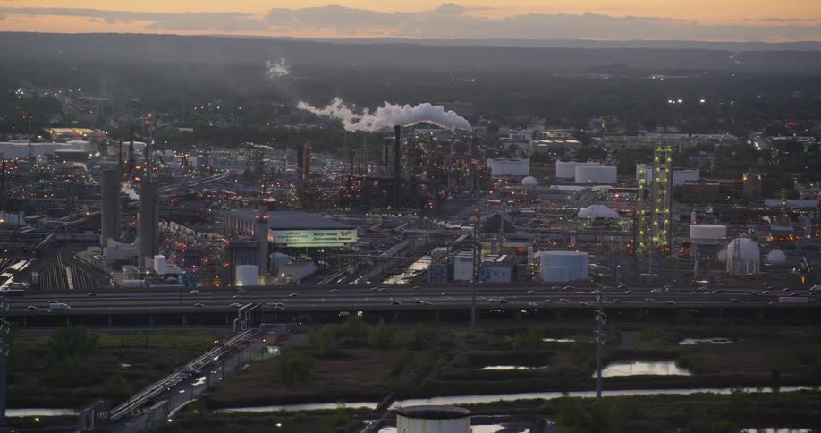 Smoke billows from the smokestack of a large factory complex next to a freeway in a city at sunset