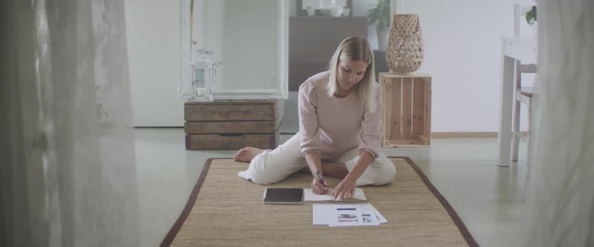A woman sits on the floor writing on a piece of paper with a tablet beside her