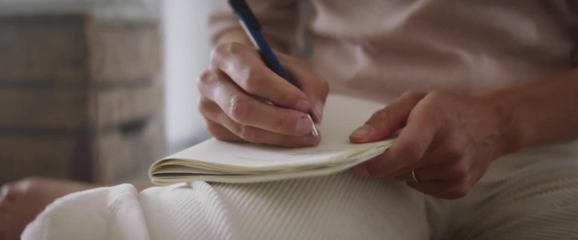 Man with a white pants and a sweater writes things down in a notebook