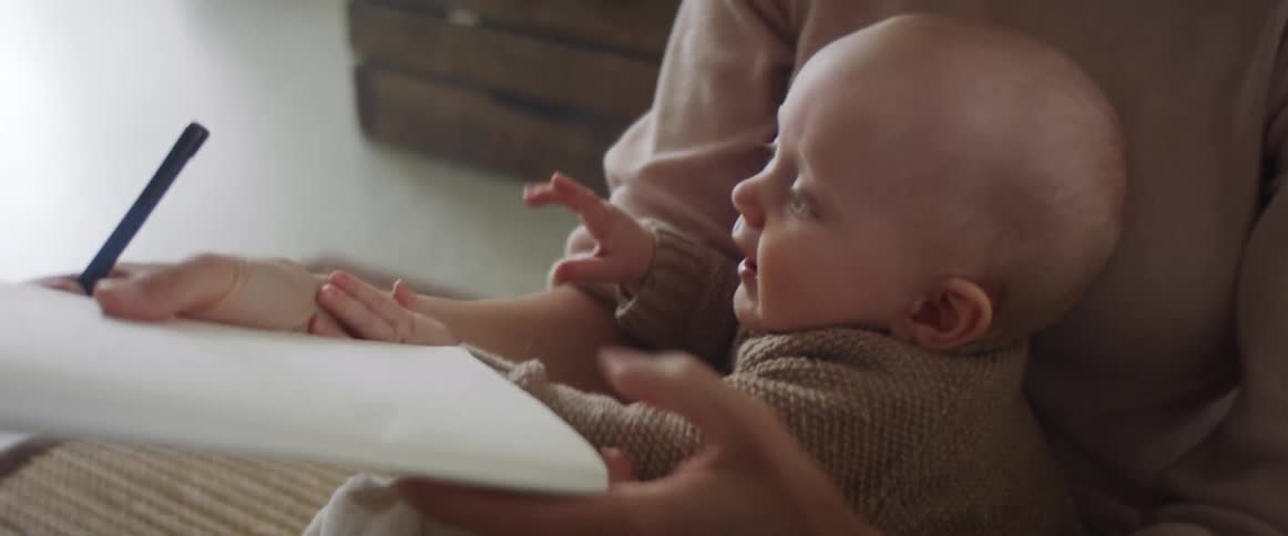 Mother holds baby on lap while sitting on the floor and trying to use a pen to write in a notebook as baby tries to grab the pen and notebook away from his mother