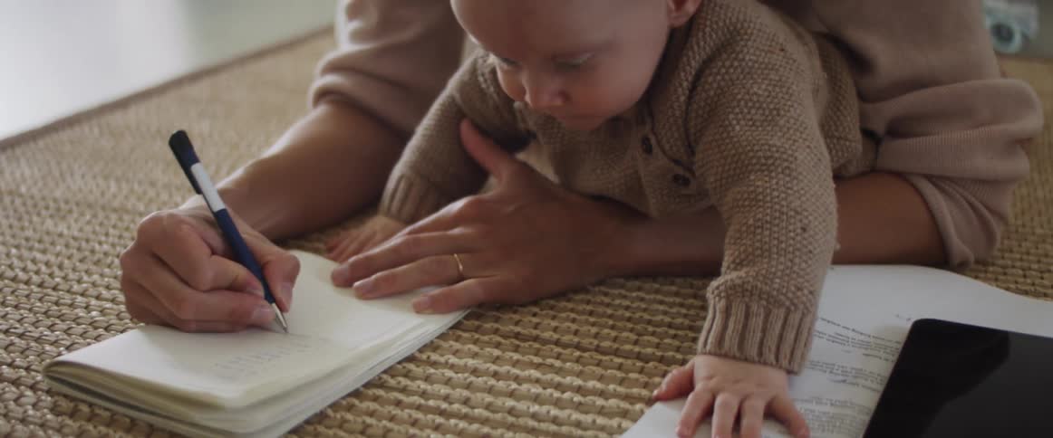 Woman holding a baby on her arms write on a notepad while lying on the floor in the living room