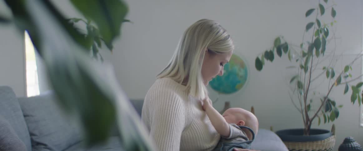A white woman with blonde hair and a tan sweater is holding her infant white son in her arms gently shushing him to sleep against her chest