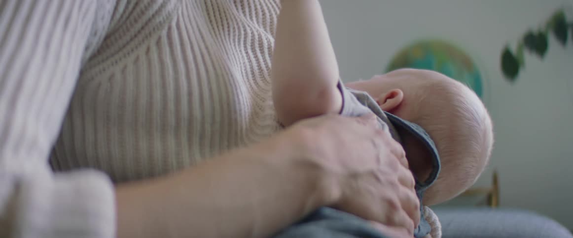 Mother with a white knit sweater sits on a sofa and holds her baby son in the living room at home