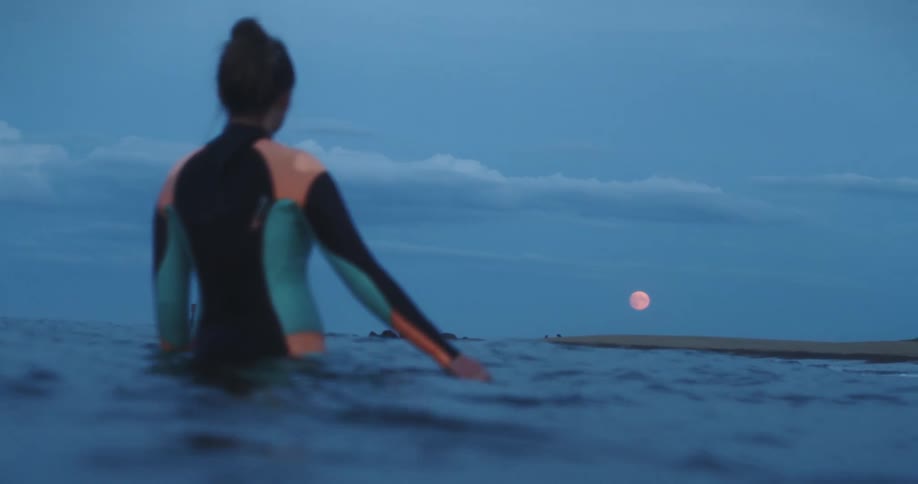 A woman in a wet suit stands in the water looking at the glowing full moon