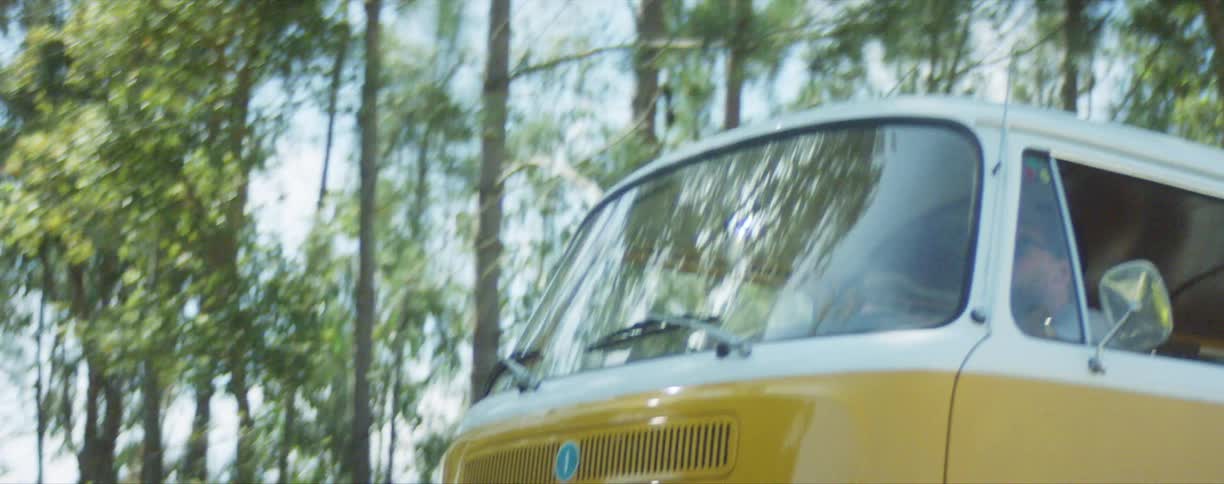 Clean yellow volkswagen van driving in the sunlight down a street with trees nearby