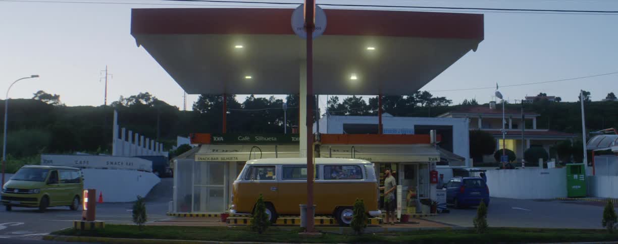 A man stands outside an orange van parked in front of a gas station at dusk
