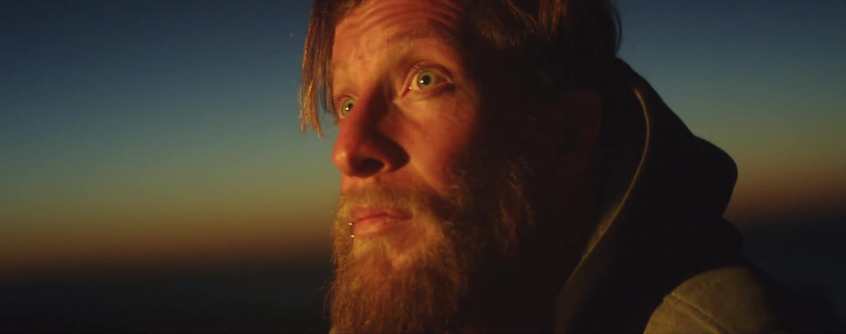 A man sits with his eyes wide open as glowing light from a campfire flashes on his face