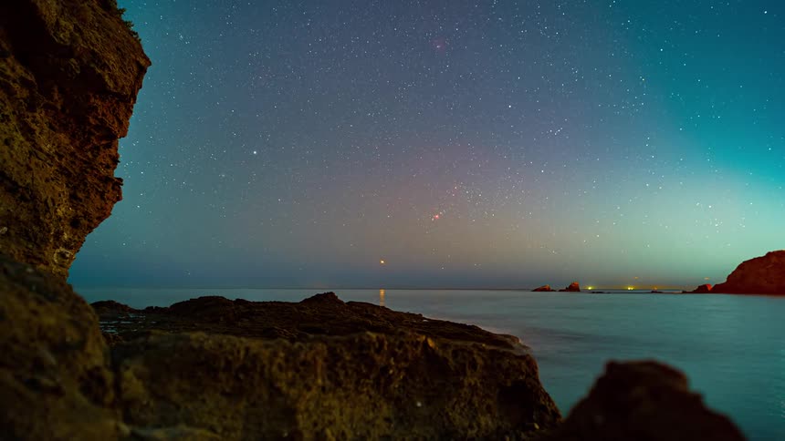 Ocean with bridge and lights in the background and stars moving in the sky