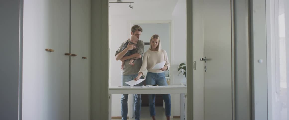 A man holding a baby stands next to a woman as she shows him papers from the desk in front of them