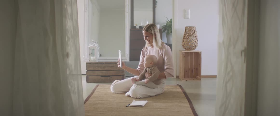 A woman sits at home on the floor with her baby in her lap and video chats on a tablet
