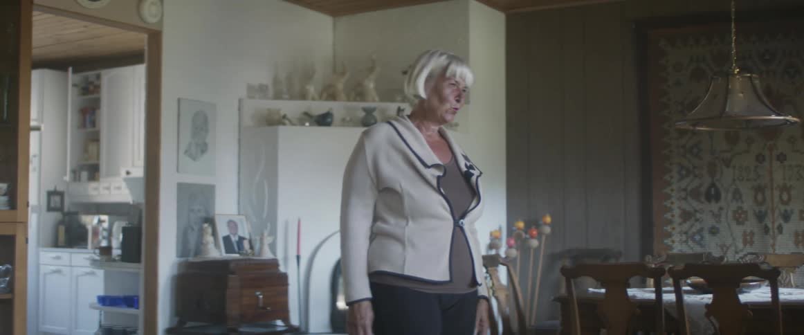 A woman stands up from an arm chair and waves at her front door before opening it and letting people in her home