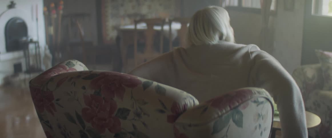 Elderly woman with grey hair sitting in an white armchair stands up and waves at those who visit her in the living room at home