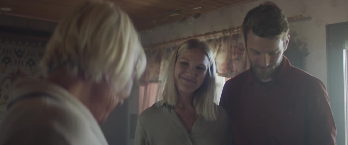 A man and woman stand together in the living room of a house as they talk to an older woman standing across from them