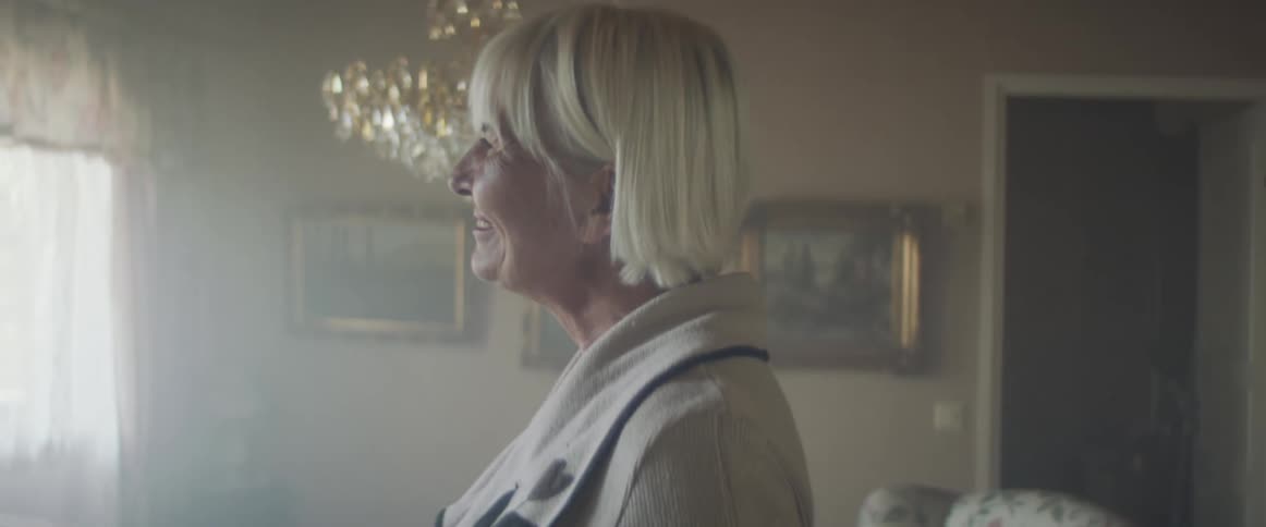 A woman stands in her living room waving outside with a smile on her face