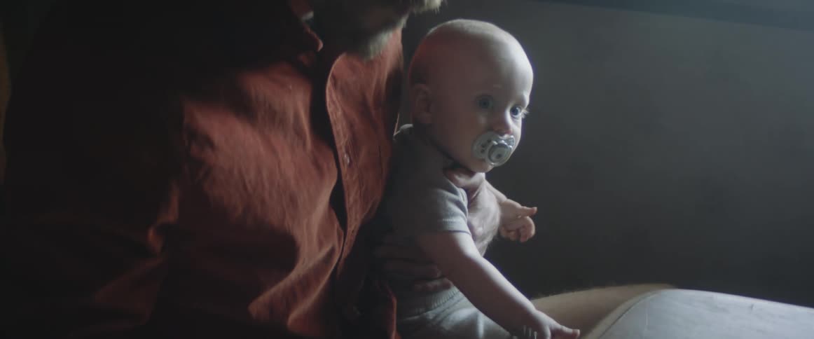 Father holds baby boy with a pacifier in its mouth on his lap securing him with one hand while he eats his meal with a fork using the other hand