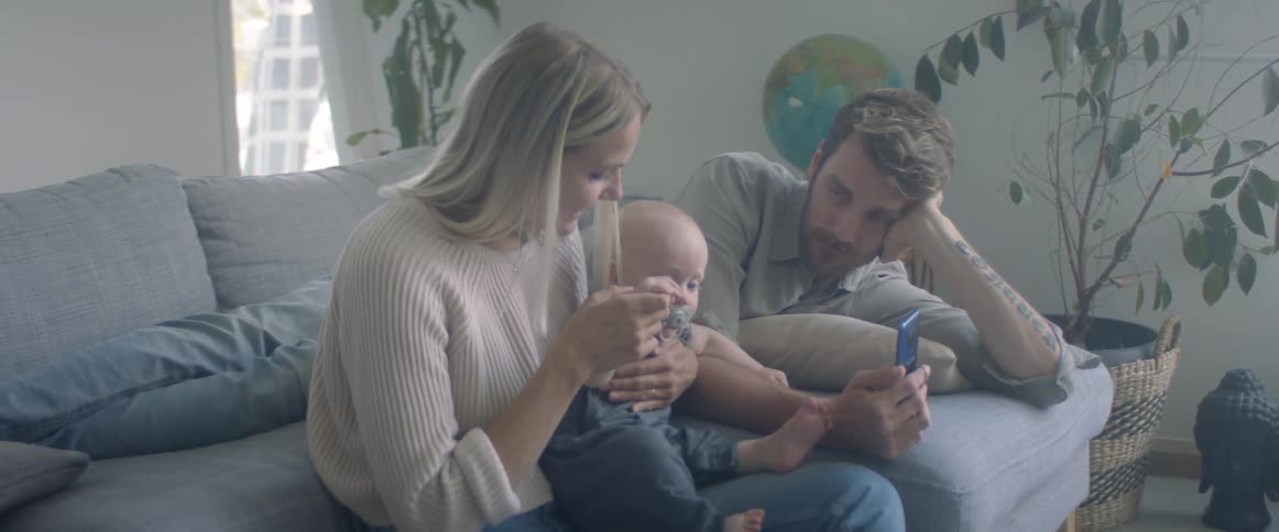 Mom waves babys hand at mobile phone screen dad is holding as they occupy couch