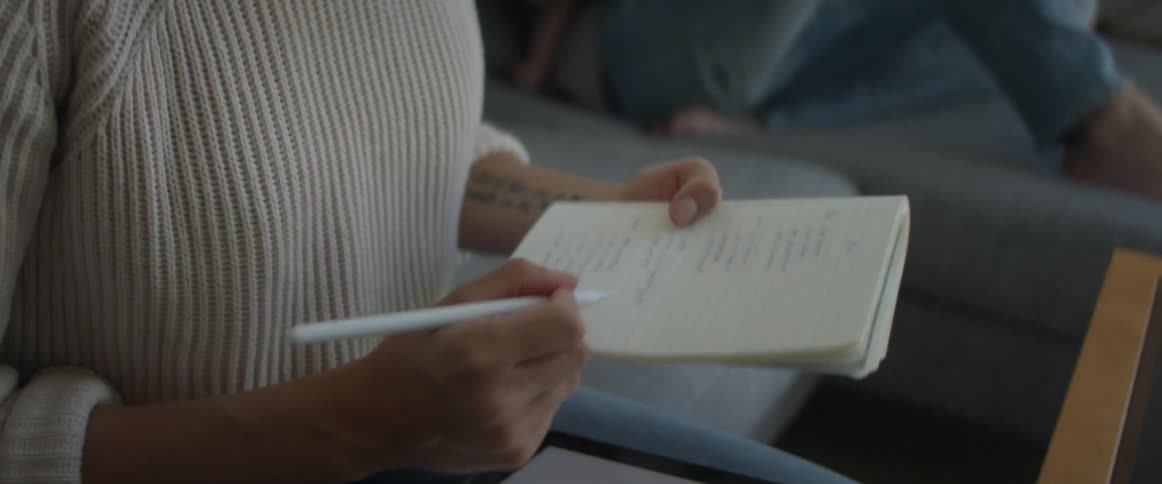 A woman writes in a notebook and on a tablet as her husband sits on the couch and holds their son