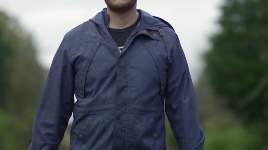 A man with a hat and jacket walking while holding a skateboard with trees the sky and clouds in the background