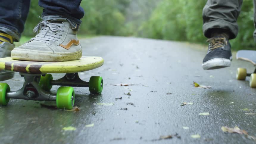 A pair of skateboarders skate side by side down a wet road on a rainy day