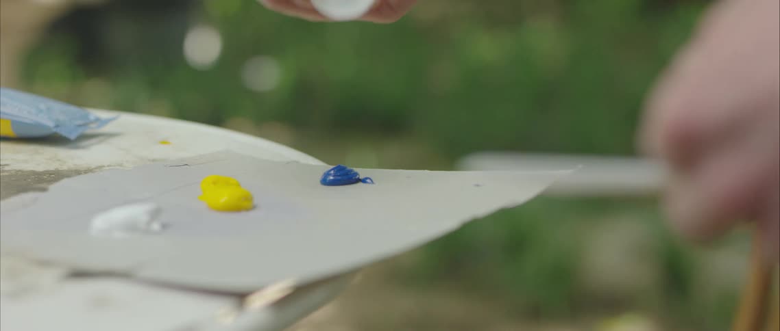 A painter adds a blob of blue paint onto a piece of cardboard being used as a palette to paint outdoors