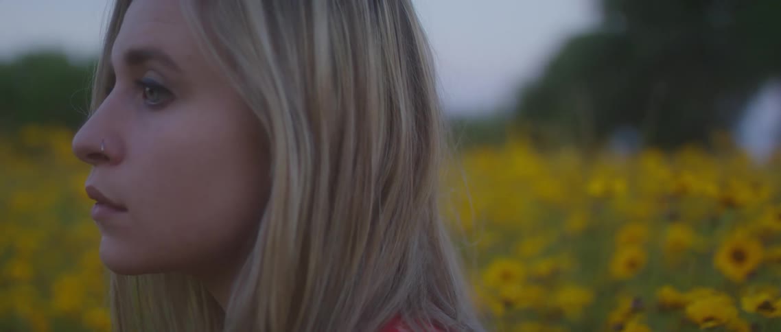 A young woman stands contemplatively in a field of yellow flowers on a cloudy evening