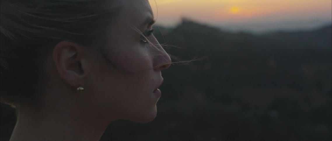 A woman standing in the mountains at sunset looks out over the countryside while wind blows through her hair
