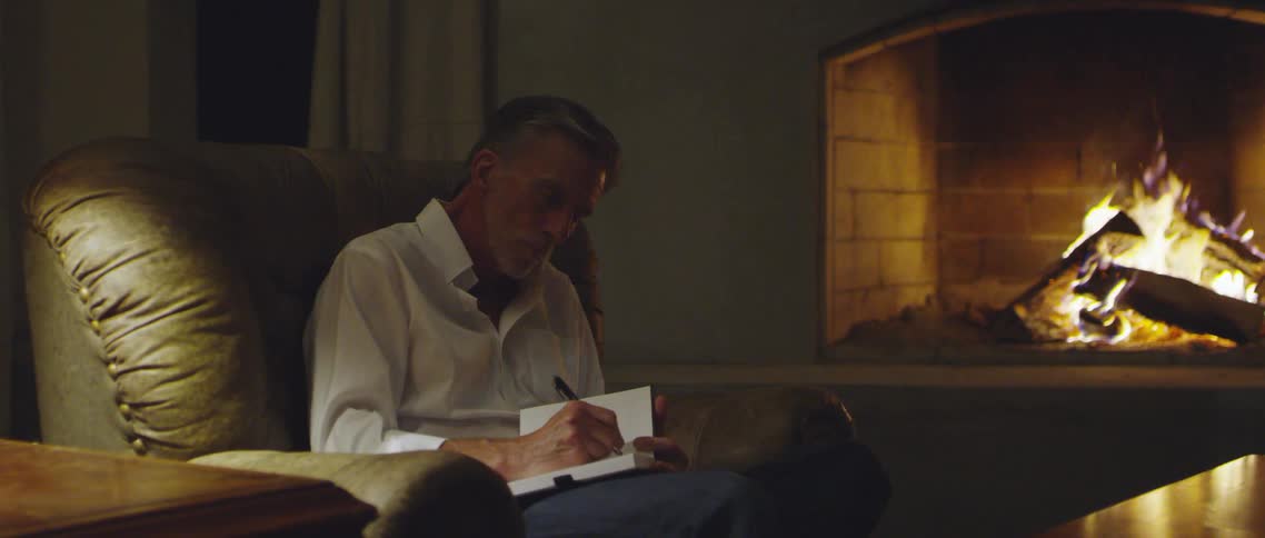 A man sitting on a couch writing on a notebook with a pen with a fireplace in the background