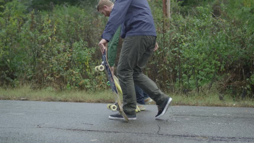 Two men walk along the shoulder of a road carrying skateboards and drop them down at the same time to skate