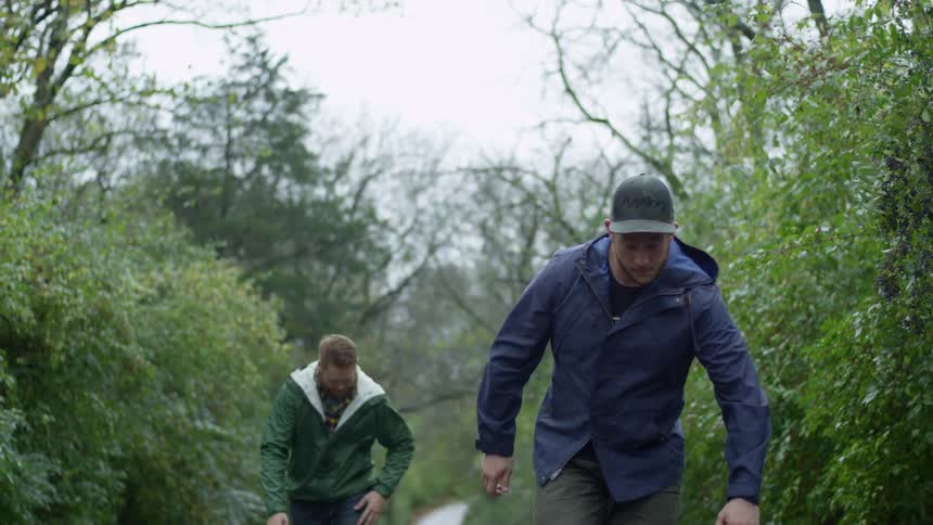 Two men skateboard along wet road through wilderness