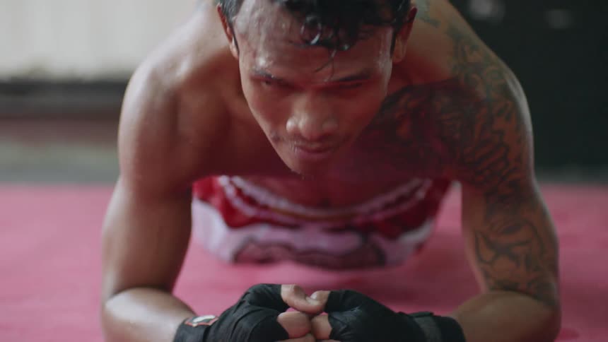 A bare chested man with black tape on his hands stretches out on a red mat in a gym setting