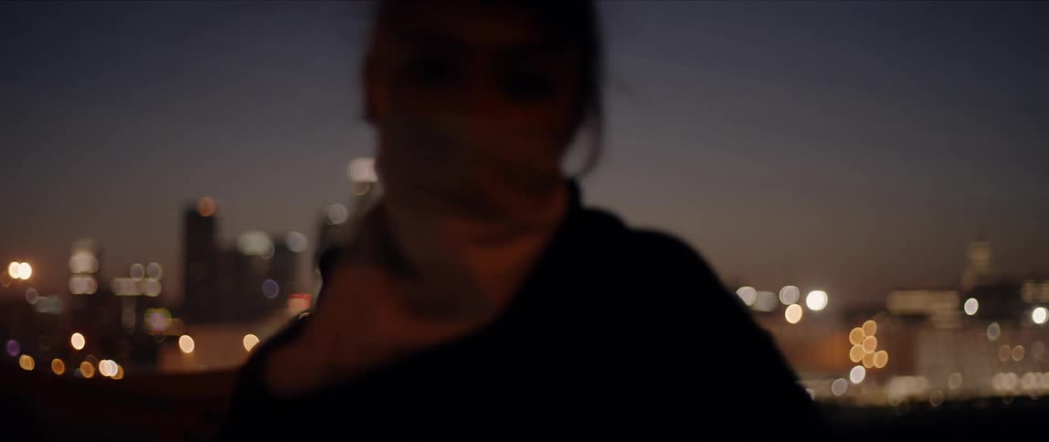 A woman with a mask on dances outside at night with the city skyline behind her