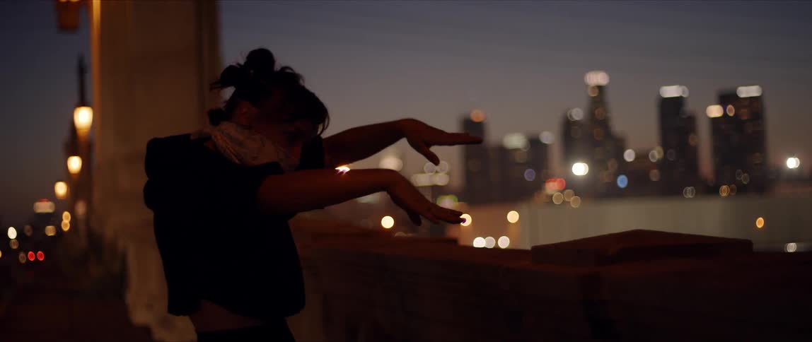 Dancer wearing bandana face covering dances on bridge lit b passing vehicle with city skyline in background