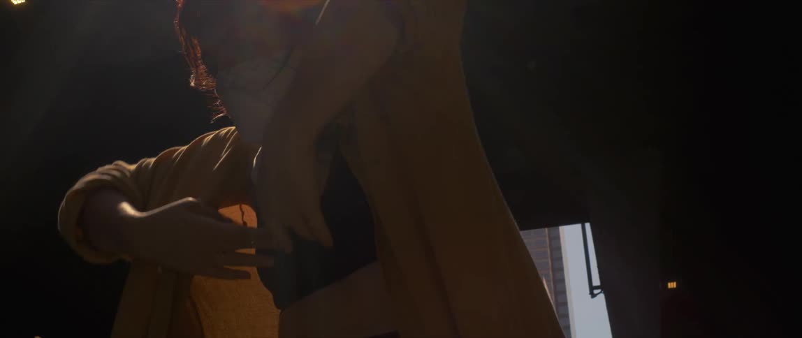 A woman wearing a scarf around her face and a yellow sweater performs dance moves inside an area that has an opening at the ceiling