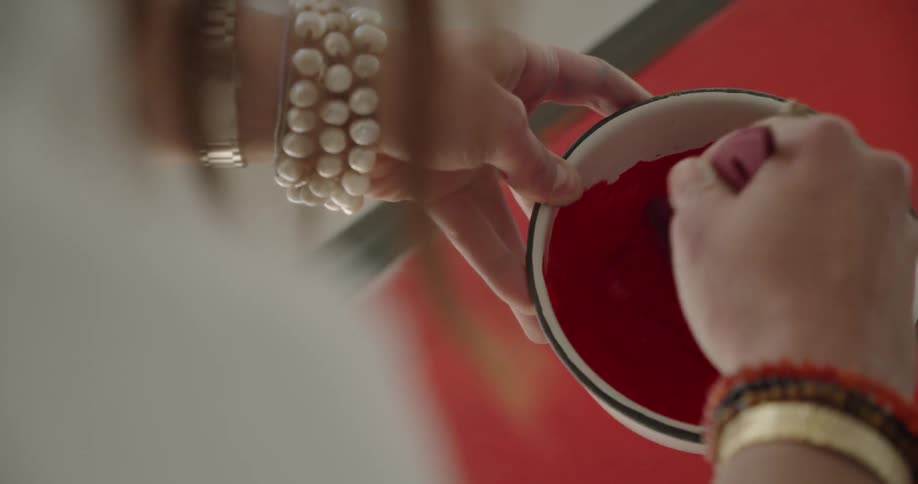 A woman wearing costume jewelry on her wrists mixes up red paint in a bowel while seated over a canvas