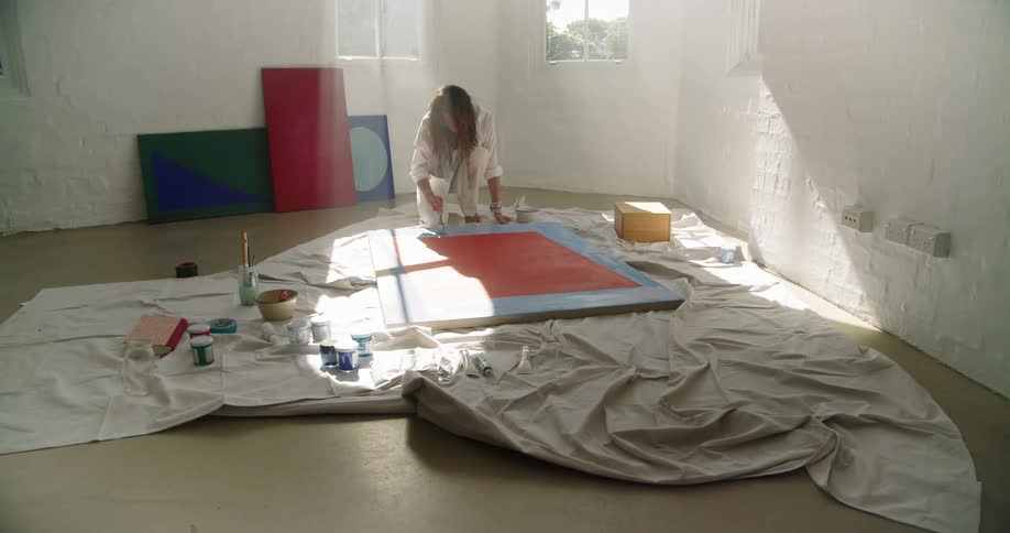 A women knells to paint a canvas that is on the floor in a room with white walls