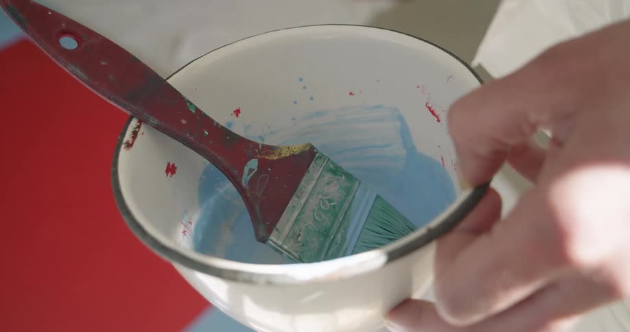A hand holding a small white bowl with a medium sized paint brush covered in light blue paint