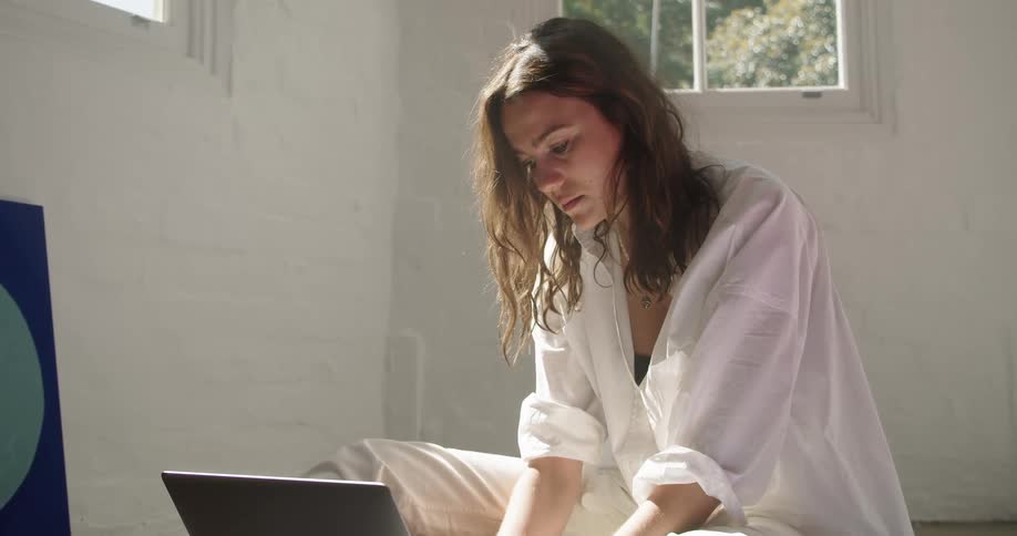 A woman sitting in a room typing on a laptop with a wall and window in the background