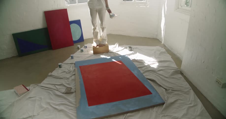 A woman in all white stands in front of a painting holding a paintbrush