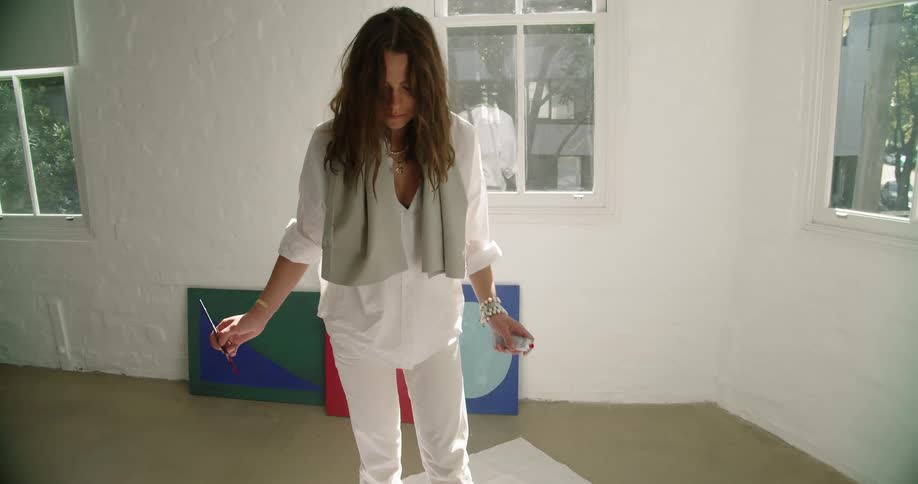 A woman wearing all white paints in orange on a canvas laying on the floor of a room