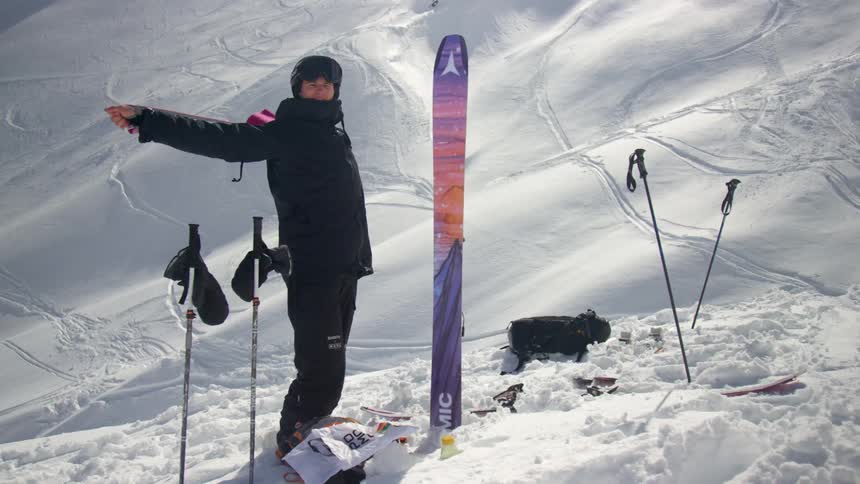 A man stands at the base of a mountain next to his skis and stretches his arms with a band