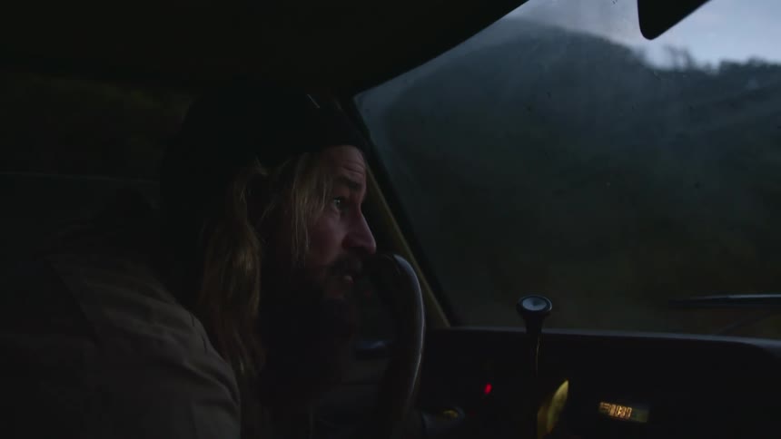 A bearded man in a vehicle leans forward to peer at something through his windshield