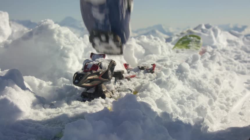 Person stomps ice crust off ski and boot with mountain on horizon