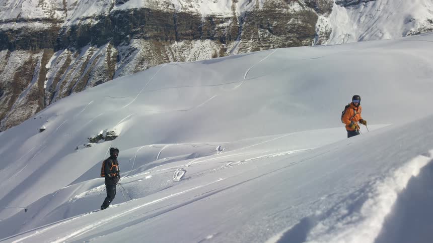 Two people with backpacks and coats standing on a snowy slope with a cliff in the background